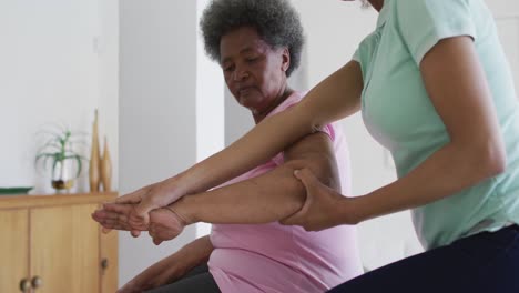 african american female physiotherapist wearing face mask helping senior female patient exercise