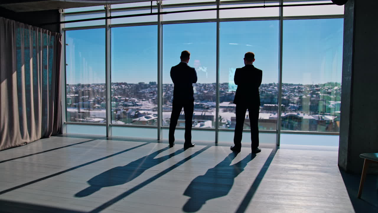 Business people talk while looking from office windows. Male business partners standing near large window indoors at sunny day. Rear view.