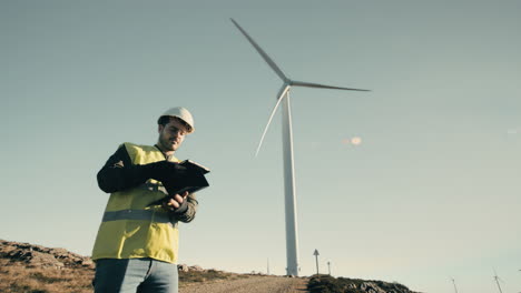 a wide shot of a young male engineer walking towards a wind turbine and then using technology on a tablet to check the windmill, emphasizing the role of innovation in the production of clean energy