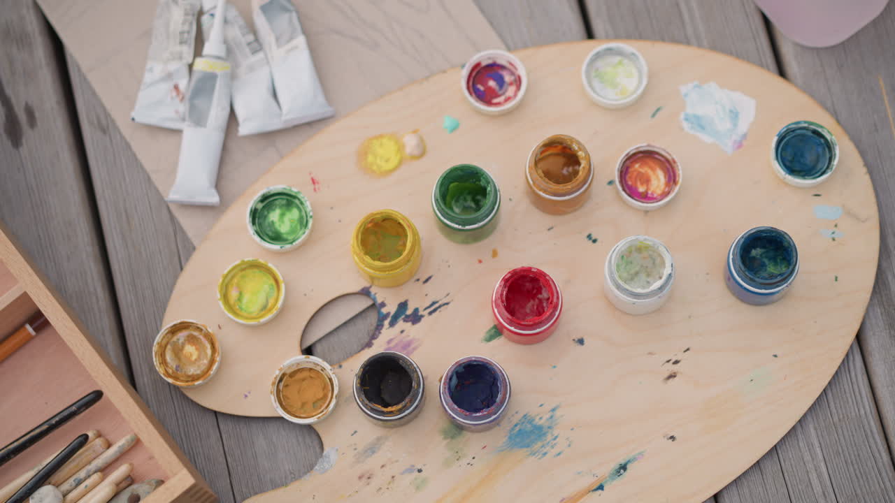palette in legno con barattoli di vernice colorati disposti su una piattaforma resistente alle intemperie accanto a pennelli e tubetti, campioni sparsi e gocciolature leggere, luce solare che evidenzia pigmenti vivaci e grana rustica, che suggerisce