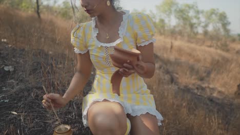 Romantic-35mm-film-look-of-Indian-woman-in-yellow-ruffle-dress,-painting-broken-pottery-shards-in-field,-shallow-depth-of-field,-smooth-slow-motion