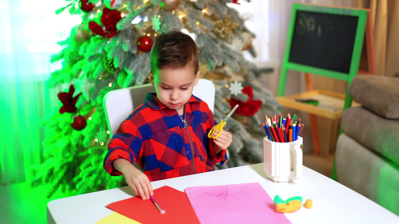 Charming toddler boy sits at desk near the Christmas tree. Little boy takes scissors and pink felt-pen