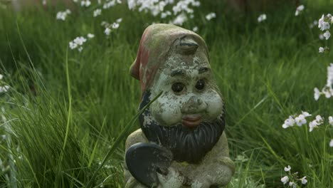 a hand places a garden gnome among tall grass and white flowers on a sunny day