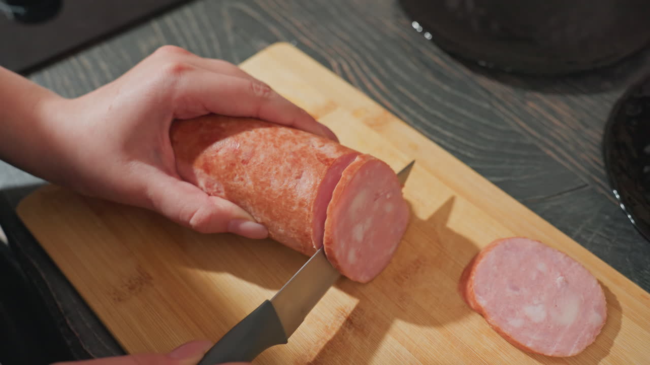 close up hands slicing pink sausage on wooden board with sharp kitchen knife during meal preparation in modern indoor kitchen showing fresh meat texture and cooking activity on dark countertop
