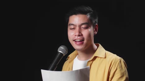 side view of asian man singer holding white paper and singing into microphone on black background