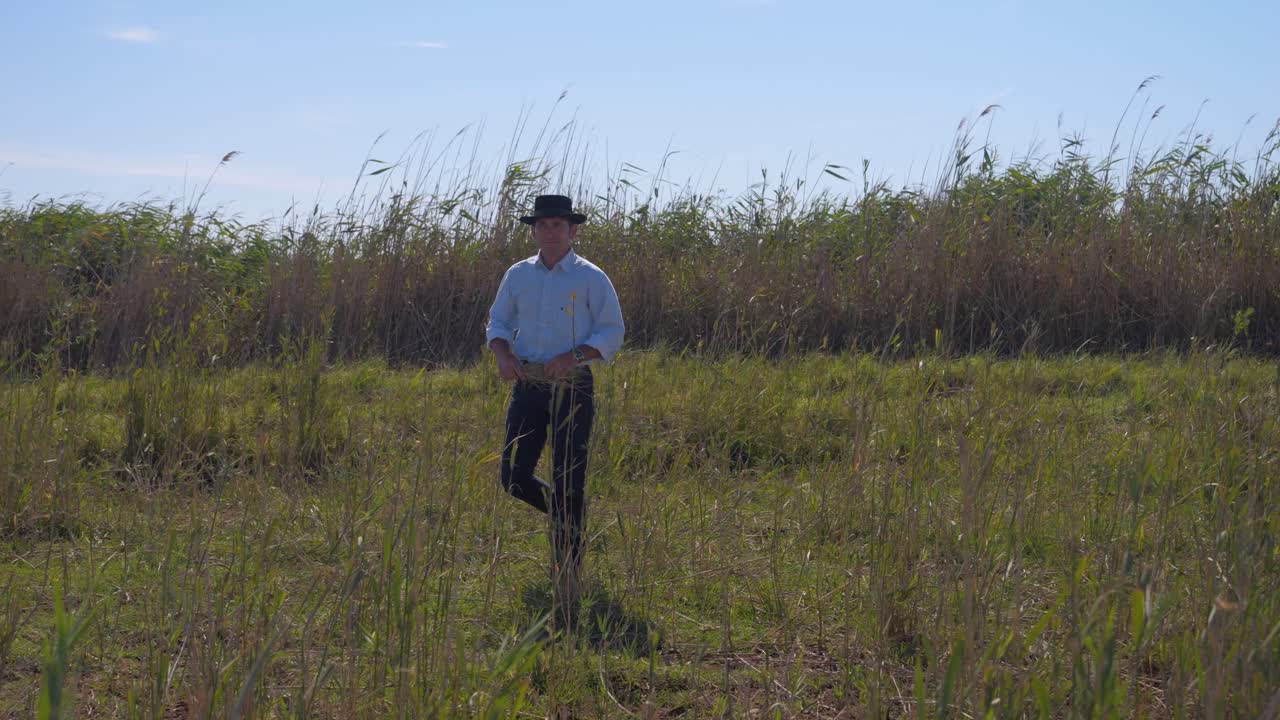 Cowboy man walking in in a field with long grass in his hand