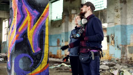 attractive girl is learning how to create graffiti under guidance of professional artist handsome bearded guy. young people are talking and laughing painting with spray paint.