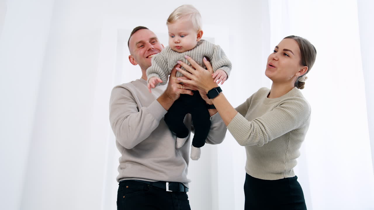 Smiling Caucasian couple holding a little baby from two sides. Parents wave their infant son. White backdrop.