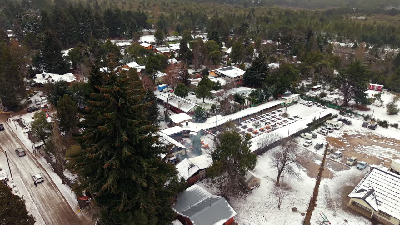 Low drone shot weaves over pines, capturing village life with chimney smoke, parked cars, and a snow-swept road. Ideal for alpine travel or winter culture reels