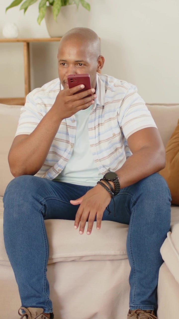 Video verticale: Un uomo con camicia a righe e orologio tiene un telefono bordeaux alla bocca e parla a casa.