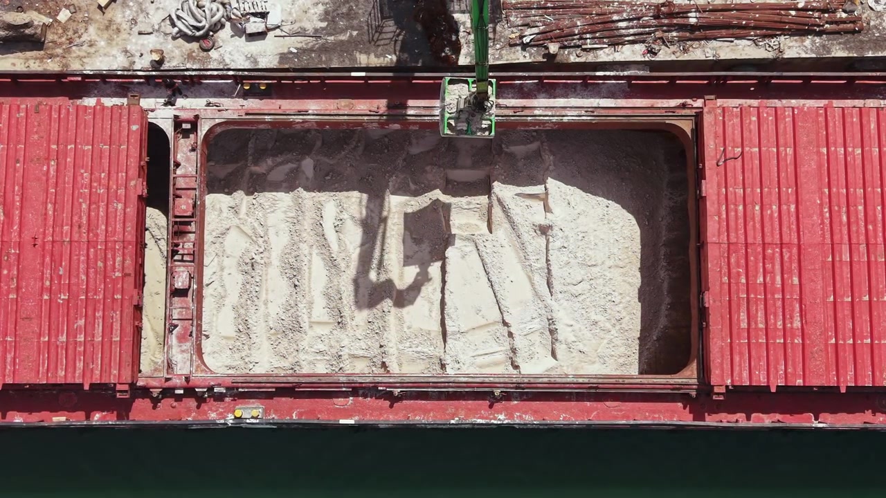 Un operatore usa una gru per scaricare il carico da una nave rossa in banchina di giorno, in un porto trafficato con container di spedizione nelle vicinanze.