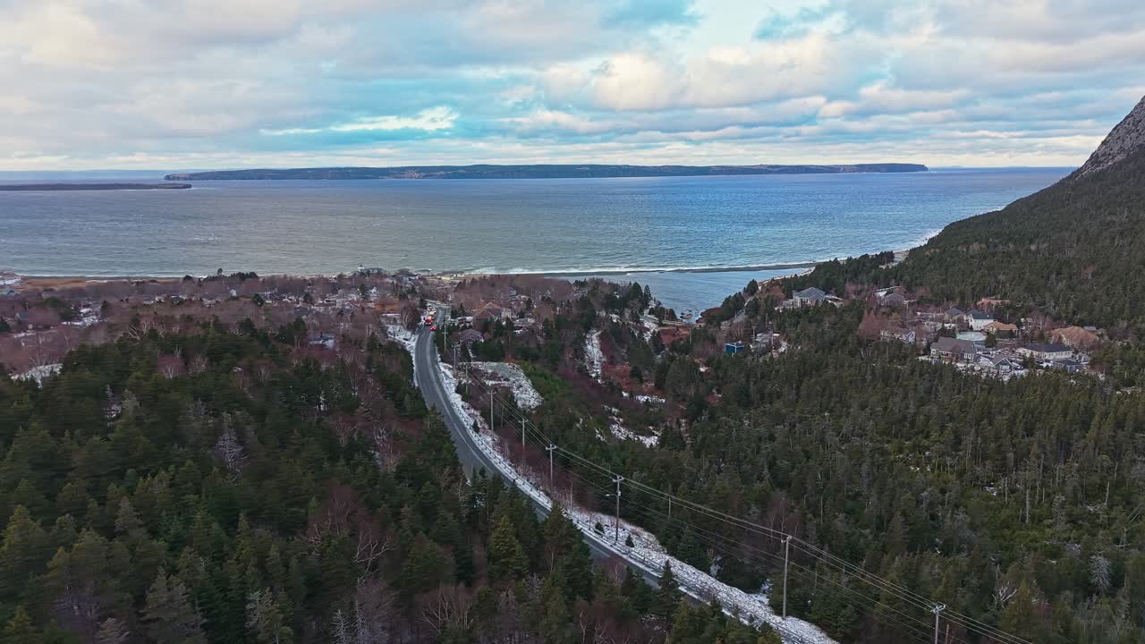Droneopptak i hpyde viser en robust skogkledd fjellside og kystbukt med sm hus plassert blant trr under en skyet himmel