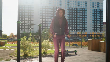 female athlete in hoodie and vest runs on spot raising arms and legs outdoors against urban backdrop under sunlight near fitness bars in park as part of warm up routine focusing on energy
