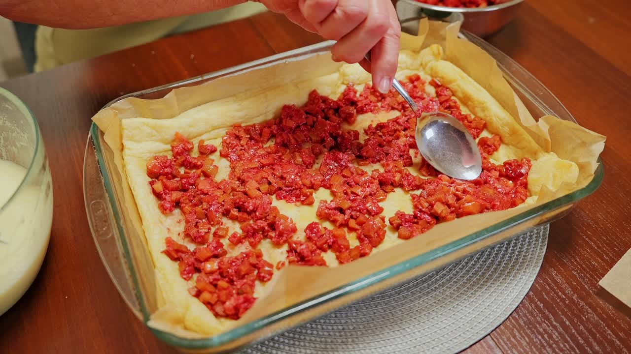 Mani che stendono uniformemente il ripieno di rabarbaro sulla pasta morbida nella teglia da forno