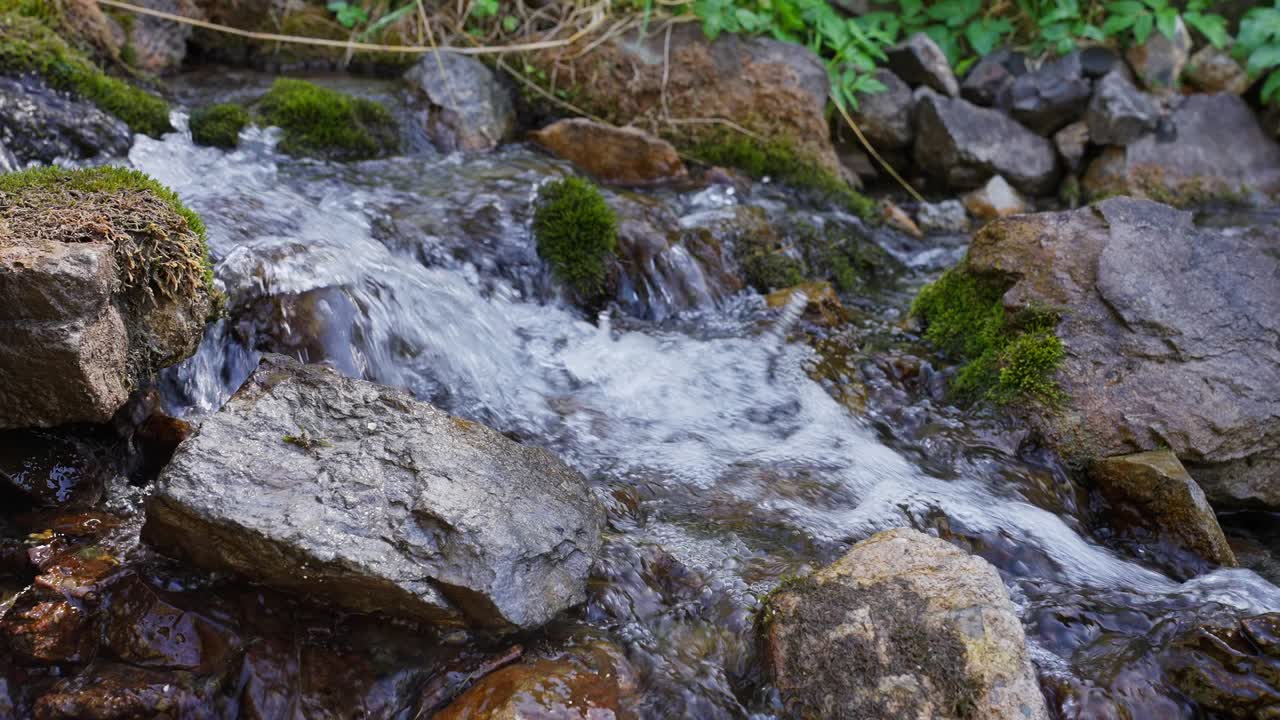 Calm mountain stream flows over mossy rocks. Water splashes gently, creating soothing white foam. Greenery frames the scene, highlighting nature's serenity. Ideal for relaxation, meditation