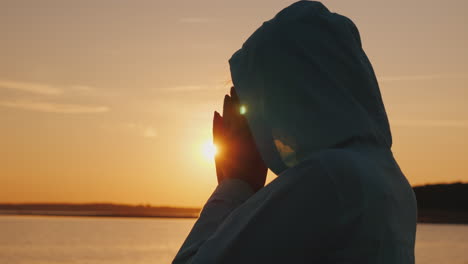 a middle-aged woman in a hood praying near the lake at sunset