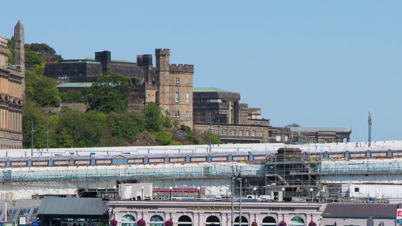 Yellow train enters busy city station under bright daylight, with historic buildings and scaffolding visible