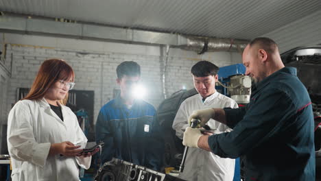 mechanic demonstrates how to loosen a bolt while students in white coats attentively nod and take notes in an automotive workshop, background includes tools, machinery, and industrial equipment