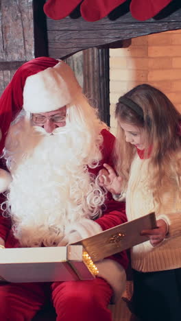 santa is reading a book for a boy and a girl near a fireplace with a fire and a christmas tree. vertical video