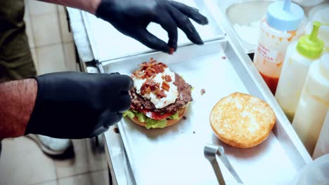 Imágenes-Cinematográficas-De-Cocina-De-Comida-De-4k-De-Un-Chef-Preparando-Y-Haciendo-Una-Deliciosa-Hamburguesa-Casera-En-La-Cocina-De-Un-Restaurante-En-Cámara-Lenta-Poniendo-Los-Ingredientes-Encima-De-La-Hamburguesa-Aplastada