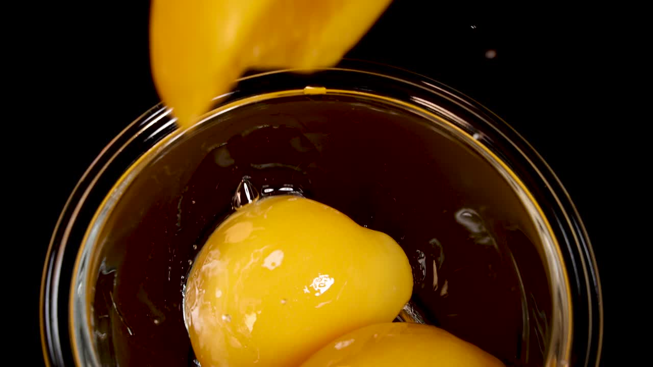 A sliced peach falls into a clear glass bowl with syrup, captured in slow motion against a dark background with dramatic, high-contrast lighting
