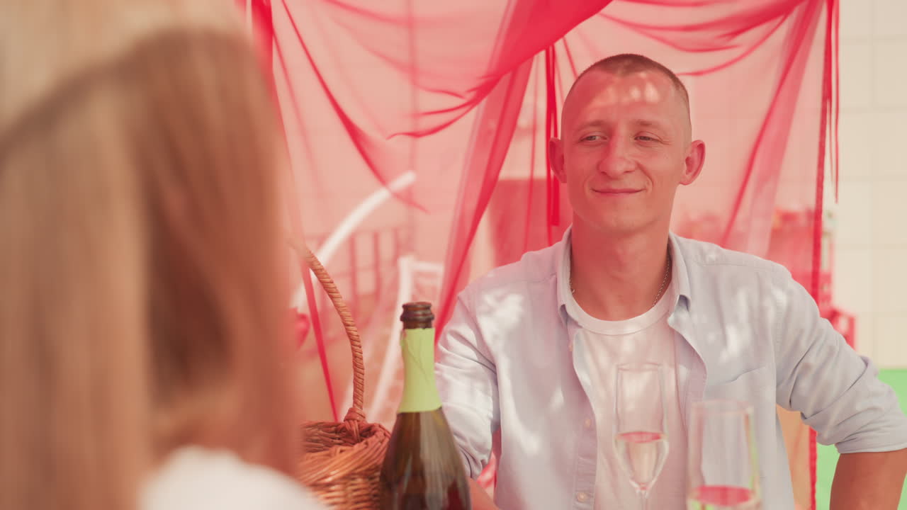 uomo bianco che offre un accendino a una donna a un tavolo da picnic, scena di appuntamento romantico con bottiglia di vino e bicchieri, gesto giocoso e sorriso invitante, donna di spalle, drappeggio rosso sullo sfondo, estate rilassante