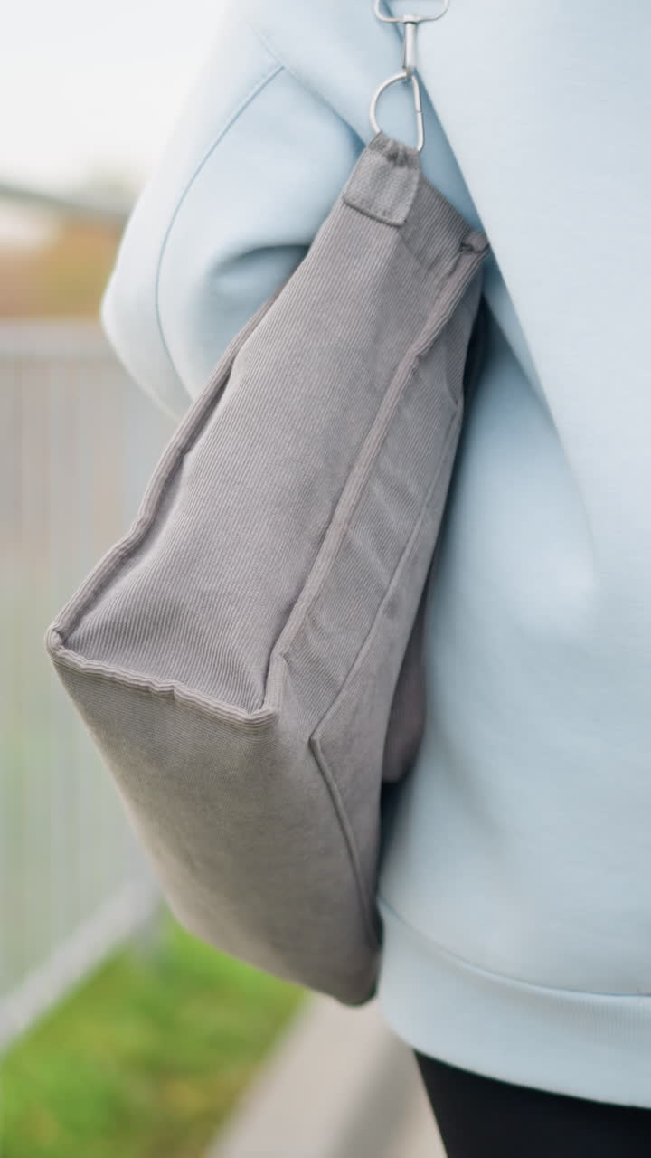 Partial view of young woman in casual wear walking with bag, with a blurred background featuring street light poles, trees, and an iron fence in a peaceful urban setting