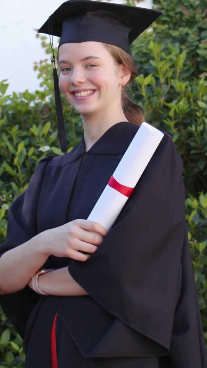 Video verticale: Registrazione, laureato che tiene in mano il diploma e sorride in giardino, indossando toga e tocco, festeggiando