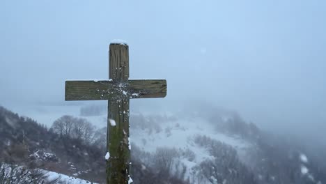 snow falling gently on a wooden cross atop a mountain, while wind sweeps snow through a serene valley below