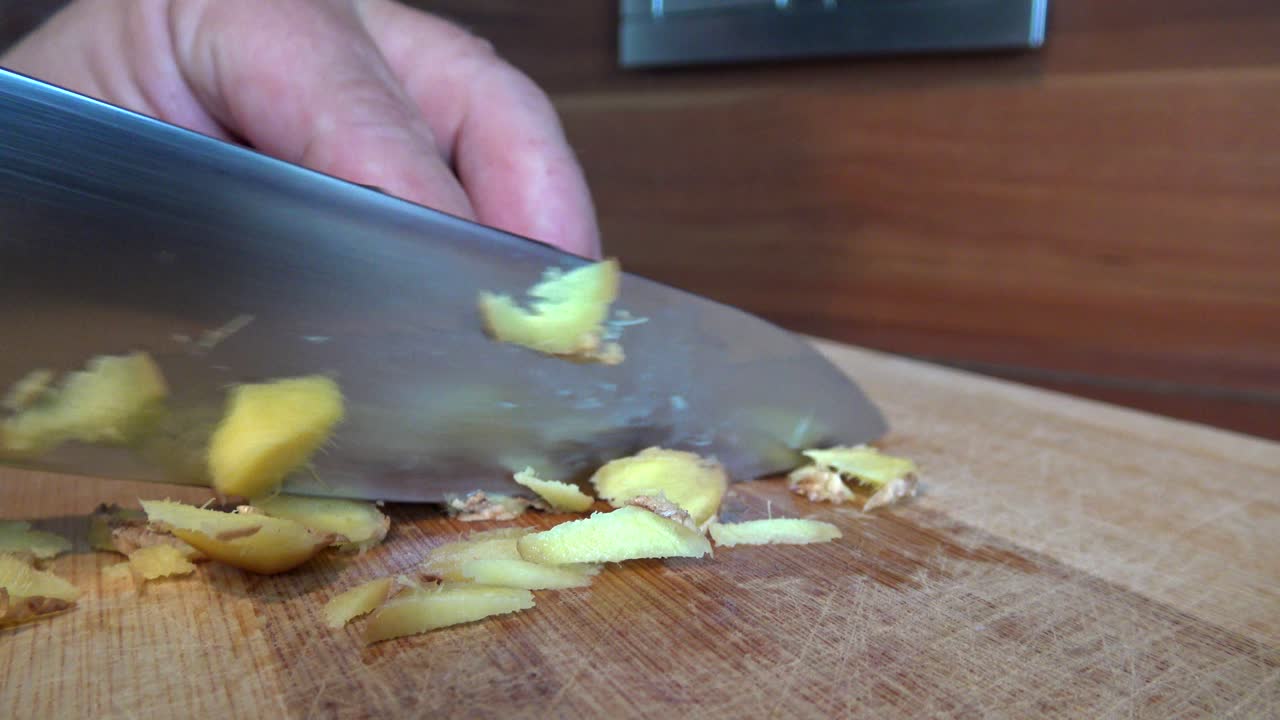 Pieces of ginger being cut into smaller pieces with a big knife on a wooden cutting board
