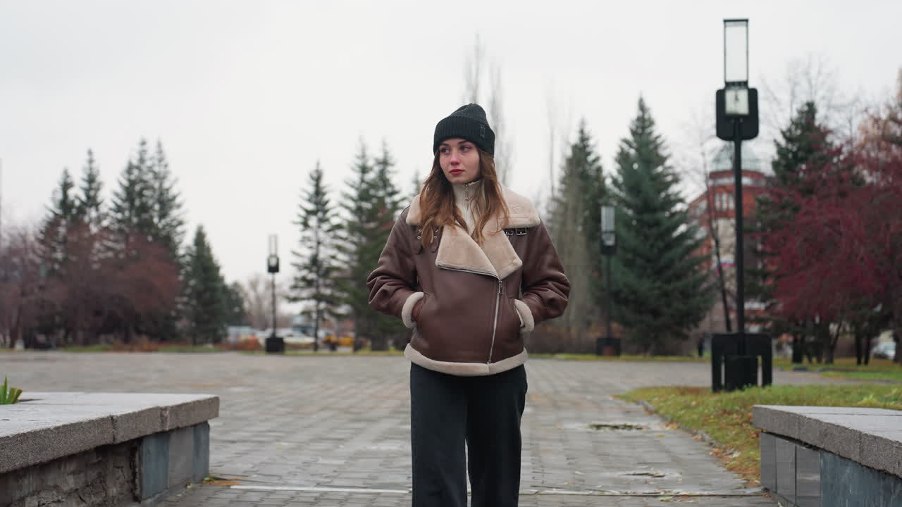 Portrait shot of young woman in black cap brown jacket walking slowly with hands in pockets on wide stone pathway through urban park lined with pine trees modern buildings and cloudy sky