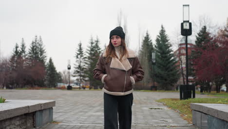 portrait shot of young woman in black cap brown jacket walking slowly with hands in pockets on wide stone pathway through urban park lined with pine trees modern buildings and cloudy sky
