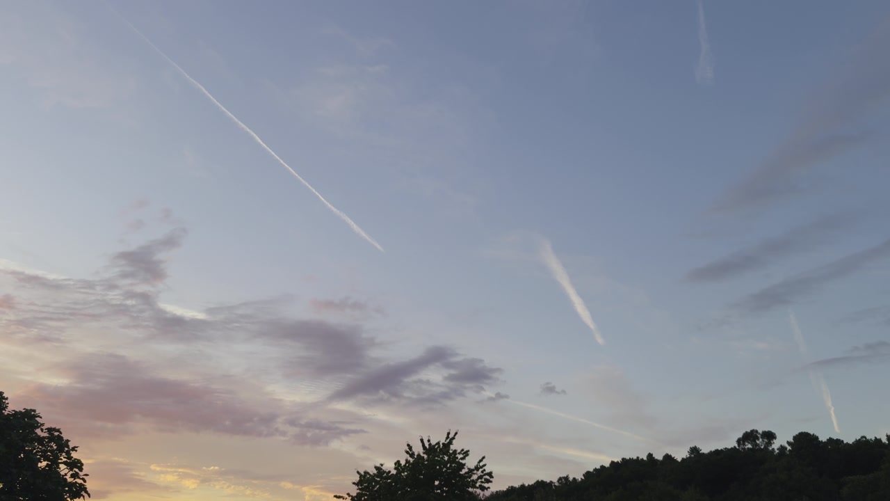 Cielo serale drammatico con nuvole color pastello e molteplici scie di condensazione di jet sopra una tranquilla collina al tramonto, ripreso con un'ampia inquadratura serena