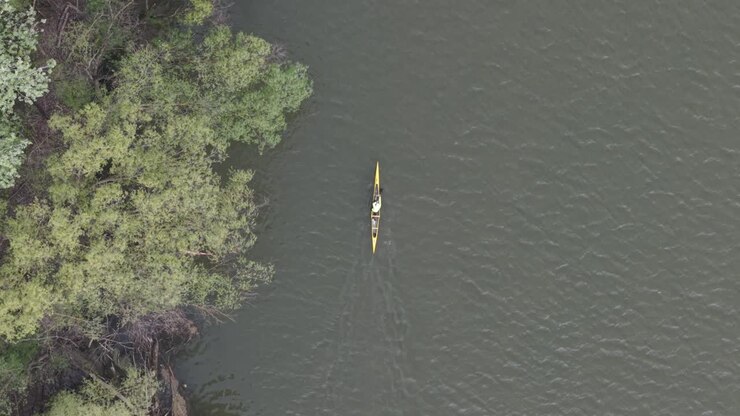 A top-down aerial view shows a lone kayaker paddling on a calm river near a green, tree-lined shore