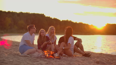 Los-Estudiantes-Están-Sentados-Alrededor-De-Una-Fogata-En-La-Playa.-Están-Hablando-Entre-Ellos-Y-Brindando-Cerveza-Al-Atardecer-Y-Disfrutando-De-La-Tarde-De-Verano-En-La-Costa-Del-Río.