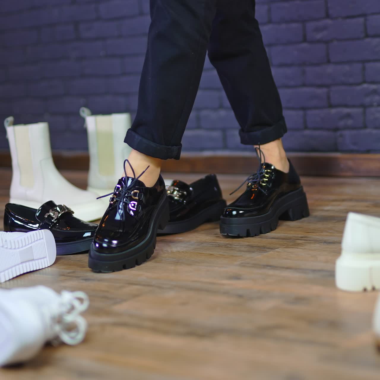 Trying on massive black shoes on tractor soles. Female feet pose among the diverse footwear scattered by the floor. Blue wall backdrop