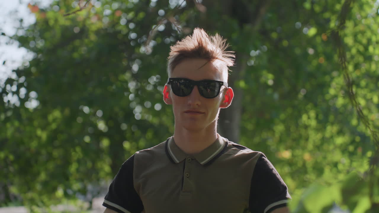 Individual Relaxing Near Trees, Person Adjusting Shades Amid Quiet Natural Surroundings Outdoors In Park, Male Individual Pauses While Wearing Sunglasses Near Trees In Serene Outdoor Park Setting