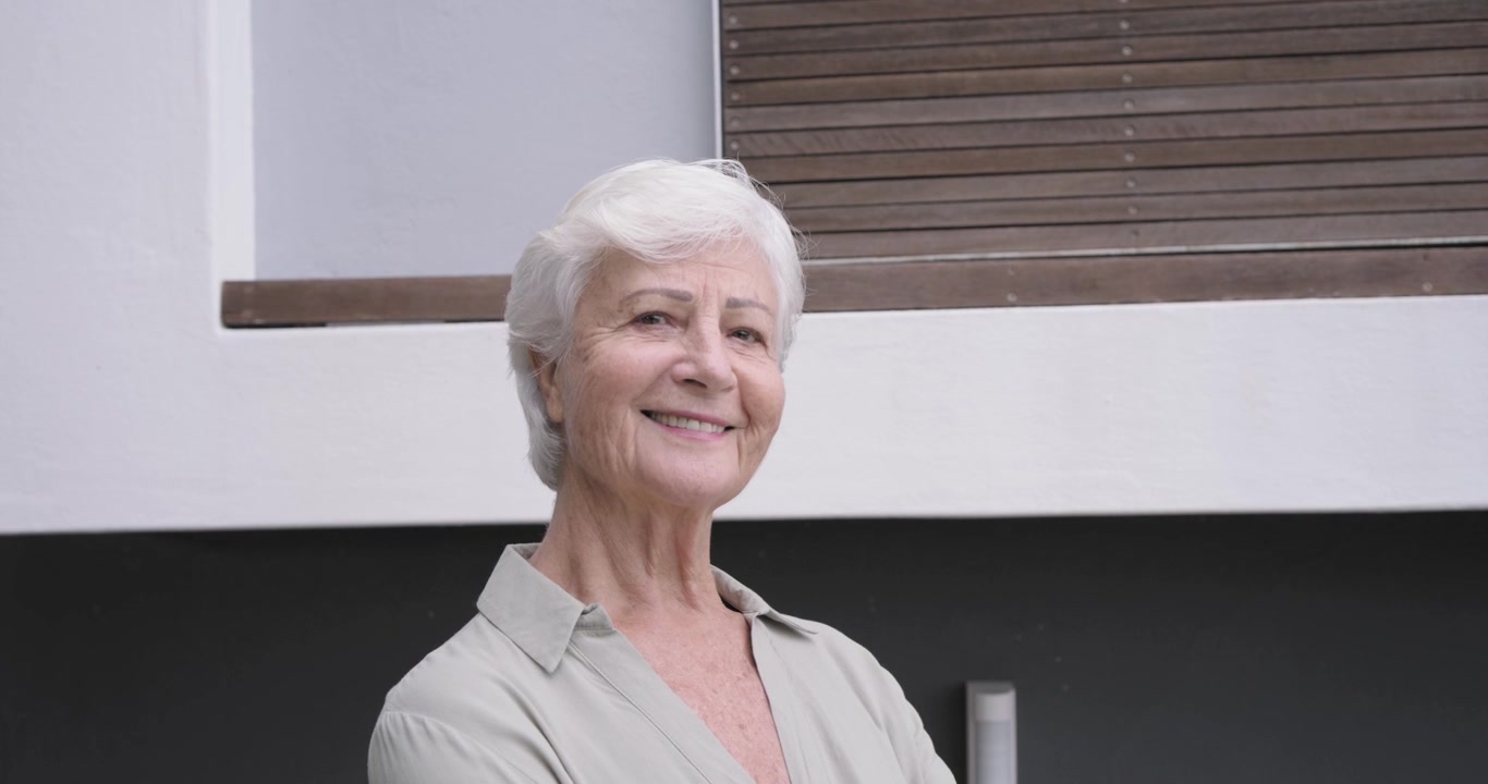 Anziana donna in piedi vicino a una ringhiera di vetro, con una camicia chiara, lo sguardo che si sposta verso l'alto mentre la telecamera si avvicina, sorridendo.