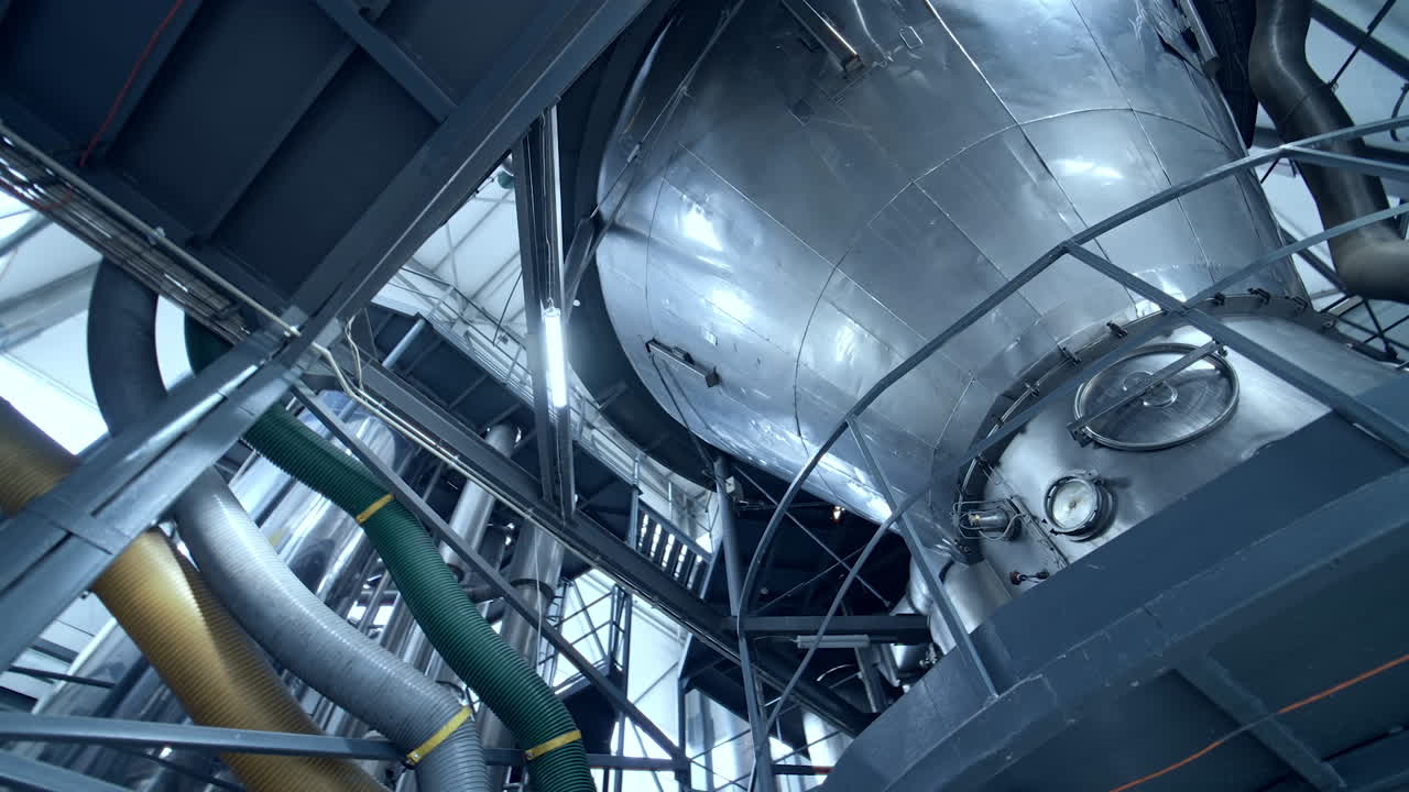 Huge dairy tank beside the thick plastic tubes. Stainless steel tank standing at the raised platform with sensors at its basics. Circle view from below.