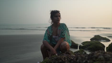 Beautiful-young-woman-sitting-down-on-mossy-rocks-on-a-sandy-tropical-beach,-at-sunset