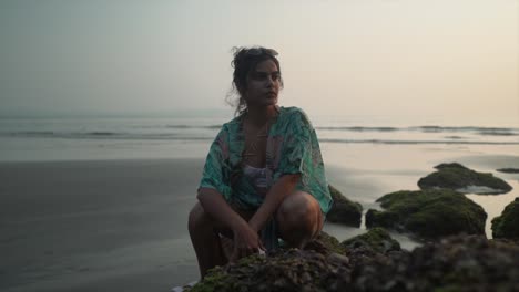 beautiful young woman sitting down on mossy rocks on a sandy tropical beach, at sunset