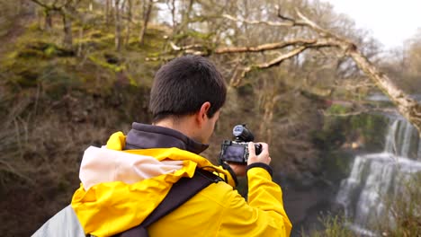 Hombre-Con-Una-Chaqueta-Amarilla-Ajustando-La-Lente-De-Su-Cámara-Para-Fotografiar-La-Cascada-Sgwd-Isaf-Clun-gwyn,-Gales