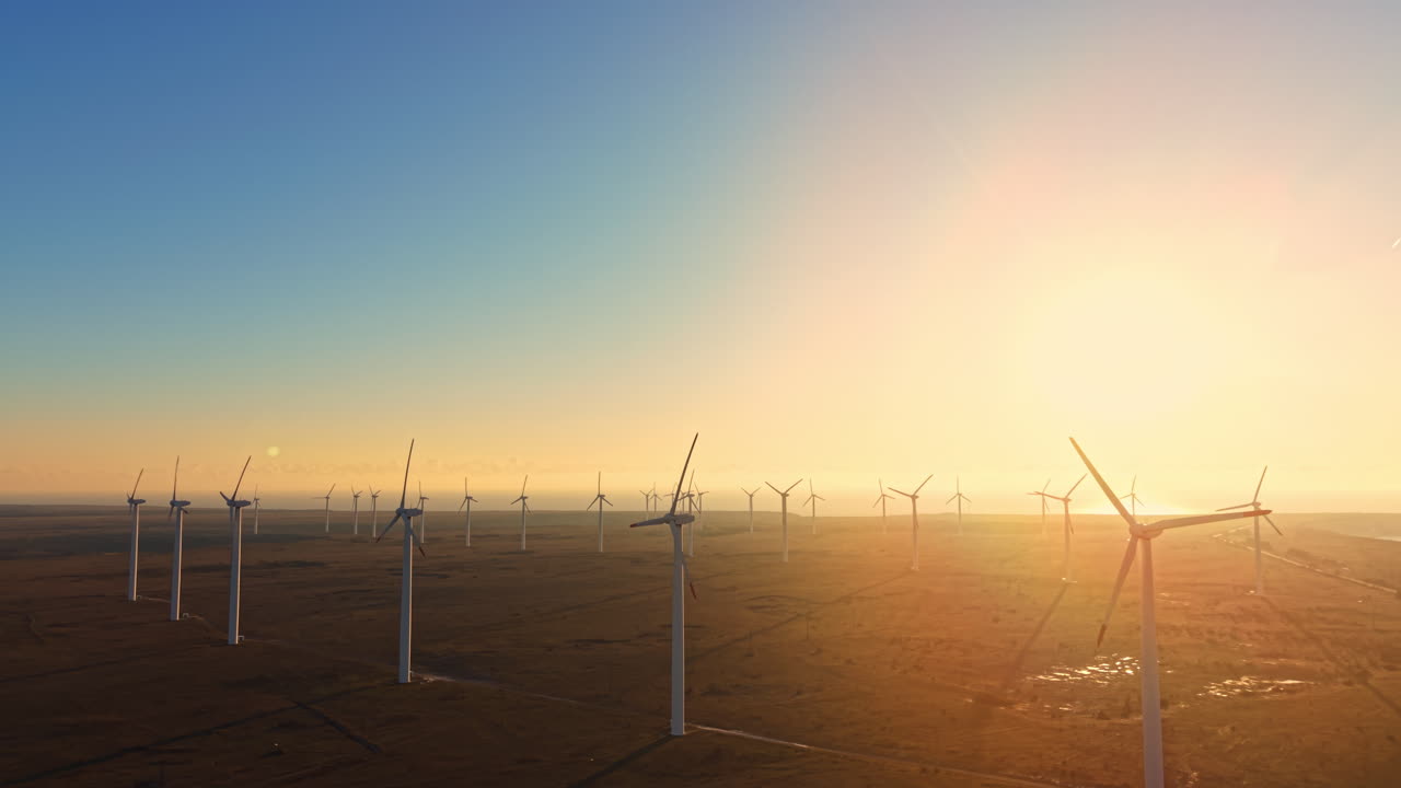 Wind turbines stand tall in a field at sunset with yellow and orange hues filling the sky and wind blowing through the blades