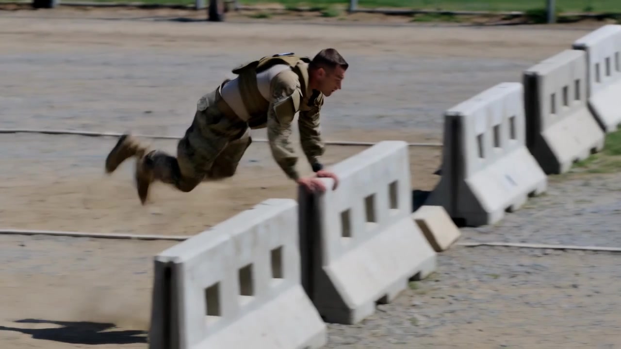 Sprinting soldier at whistle wearing tan armor vaulting barriers on training lot kicking dust