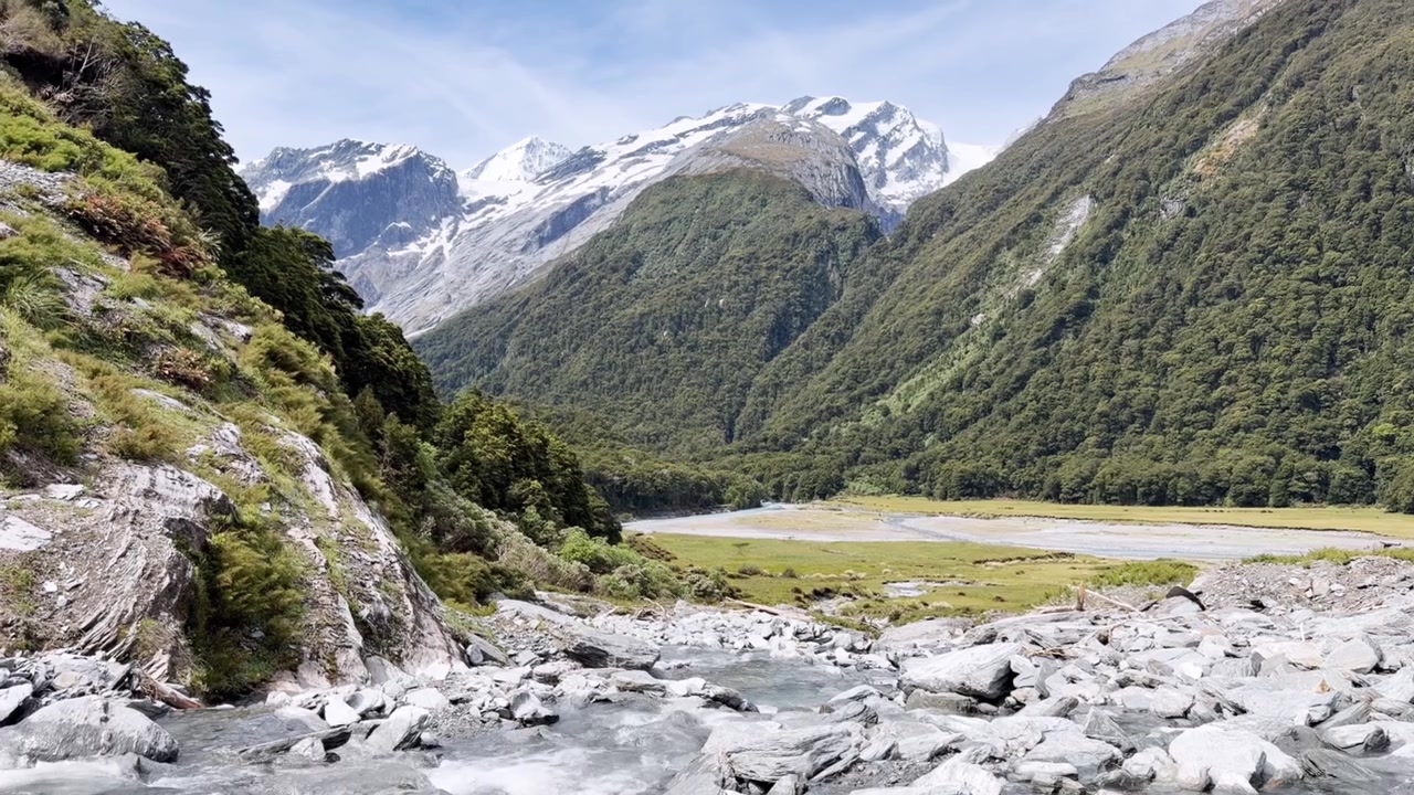 Sereno paesaggio montano con cime innevate, vegetazione autoctona e un fiume intrecciato che attraversa una valle alpina incontaminata sotto cieli azzurri e luminosi