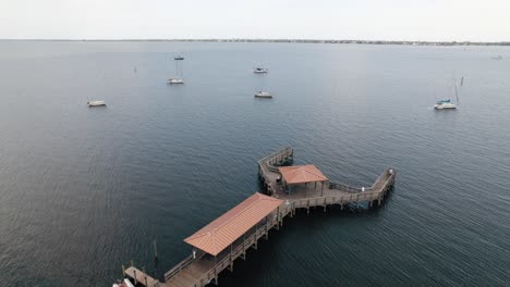 drone shot flying over pier and blue water with sailboats in the background