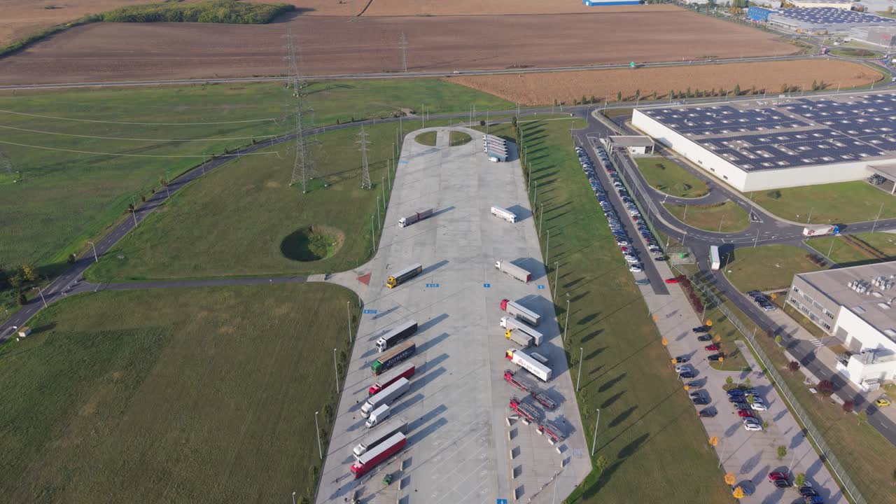 An aerial video showing a large logistics center with a wide truck parking area surrounded by green fields. Several semi-trailers and trucks are parked near the industrial warehouse under daylight