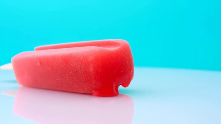 A single red popsicle rests on a glossy reflective surface against a bright turquoise backdrop, creating a clean minimal studio composition with soft lighting