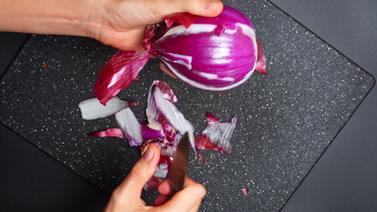 Mani che sbucciano una cipolla rossa in una cucina di giorno. Una persona usa le mani per sbucciare una cipolla rossa su un tagliere scuro in una cucina