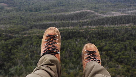 Alte-Abgenutzte-Wanderstiefel-Aus-Leder,-Die-über-Den-Rand-Der-Klippe-Hängen-Und-Sich-Langsam-Bewegen,-Während-Sich-Die-Wildnis-In-Die-Ferne-Erstreckt